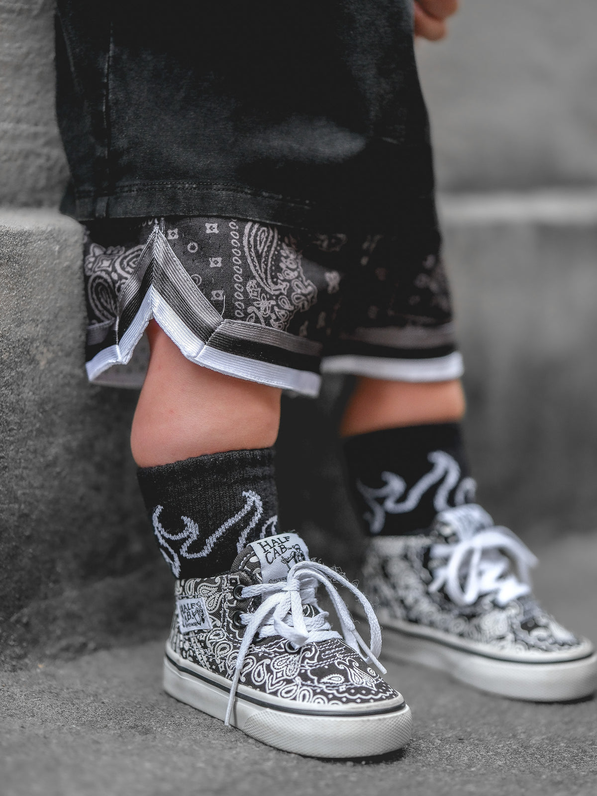A close-up lifestyle shot of a child's legs wearing black and white paisley bandana sneakers, black socks with a white graphic, and black shorts with a bandana print and stripe detail at the  hem standing on gray concrete steps.