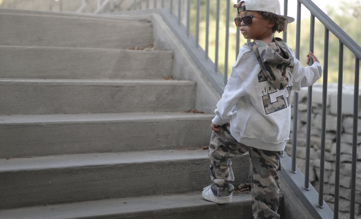 A child wearing a white hoodie with a large graphic print on the back, camouflage pants, and a white cap stands on concrete stairs next to a metal railing.