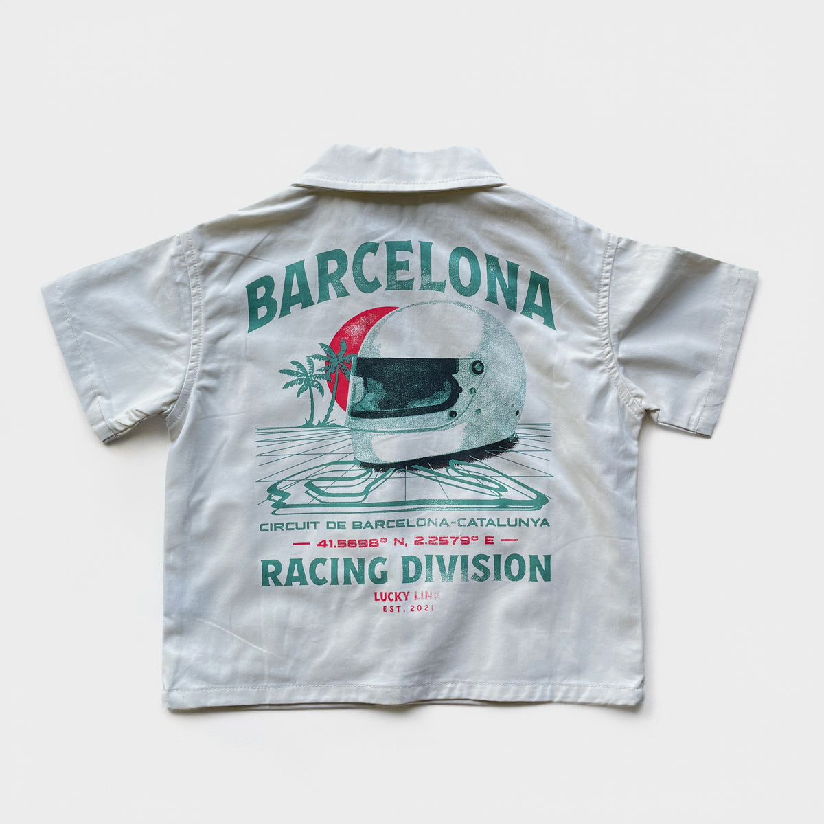 A cream short-sleeve  overshirt shown from the back, featuring a vintage-style racing graphic with the word 'BARCELONA' in teal, a white racing helmet with a red sun motif, and 'RACING DIVISION' printed below.
