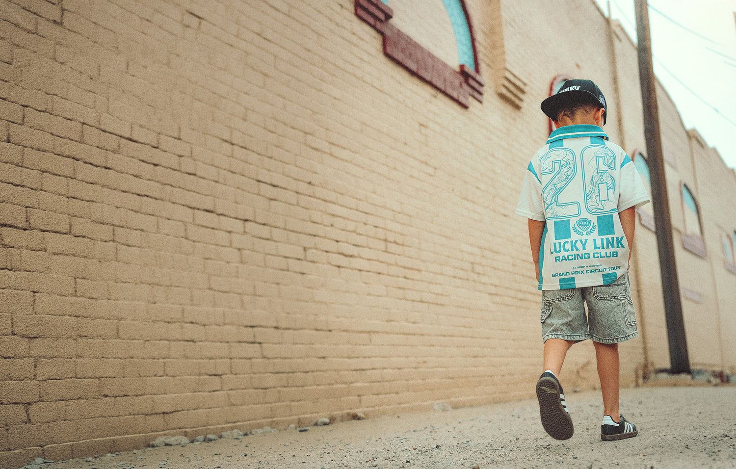 A child wearing a white and teal 'Lucky Link' racing collared jersey, denim shorts, and a black cap walks away from the camera along a sunlit dirt path next to a tan brick wall.