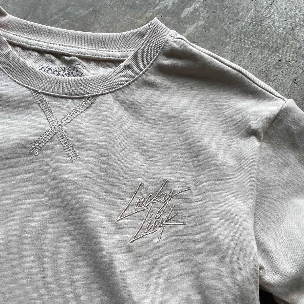 A close-up of a light beige crewneck t-shirt featuring a tonal embroidered 'Lucky Luck' script logo on the left chest and a decorative tonal cross-stitch pattern on the right chest, laid flat on a textured gray surface.