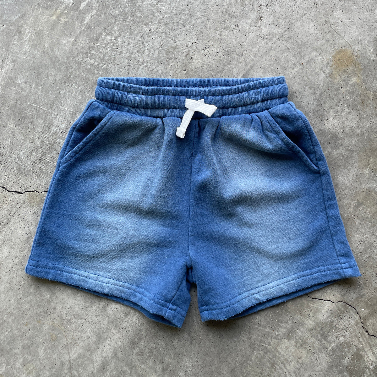 A pair of blue faded sweat shorts with a white drawstring, laid flat on a gray concrete surface.
