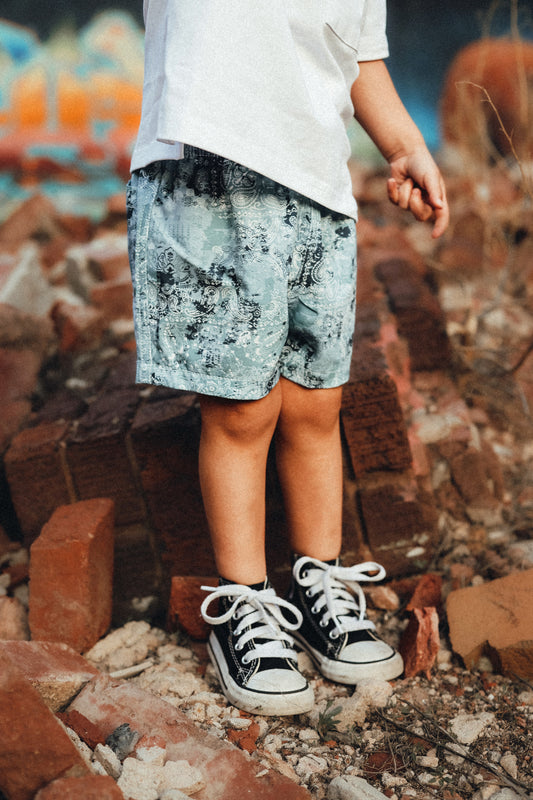 A pair of swim shorts in a blue paisley pattern with black details, worn by a child standing on a brick surface with graffiti in the background.