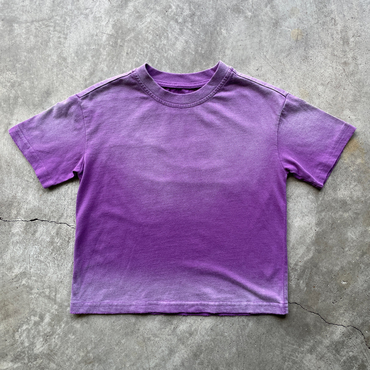 A purple short-sleeve crewneck t-shirt with a gradient fade, laid flat on a textured gray concrete surface.