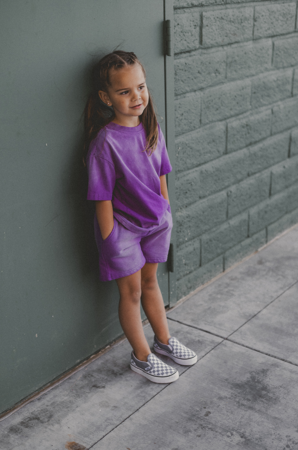 A young girl with her hair in braids stands against a dark green brick wall wearing a matching purple gradient shirt and shorts set, paired with gray and white checkered slip-on shoes.