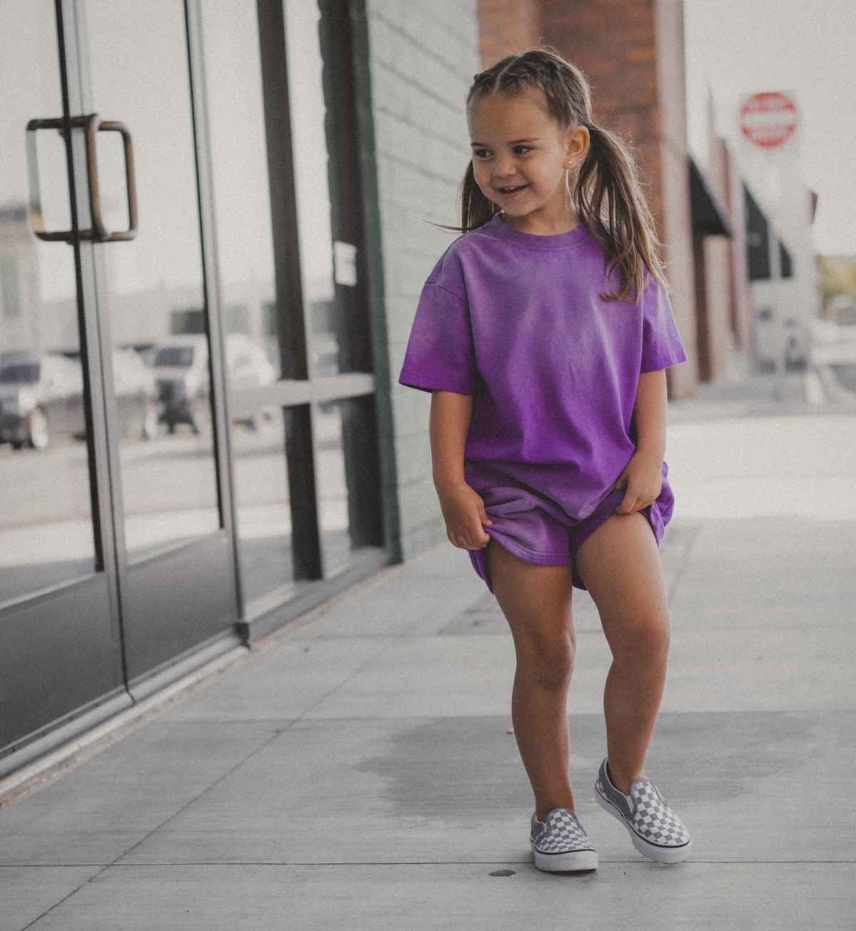 A young girl with pigtails stands on a sidewalk wearing an oversized purple t-shirt and matching purple shorts, paired with black and white slip-on shoes.